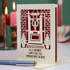 A personalised Christmas card with a laser-cut design of a front door, wreath, and small Christmas trees, displayed on a table with a pen in the foreground and lit candles in the background.