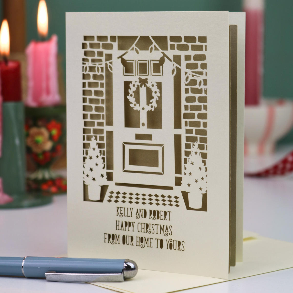 A personalized Christmas greeting card with a laser-cut illustration of a front door, wreath, brick walls, and potted trees, with the text 'KELLY AND ROBERT HAPPY CHRISTMAS FROM OUR HOME TO YOURS'. The card is standing upright on a tabletop with a blue pen in the foreground and blurred candles in the background.