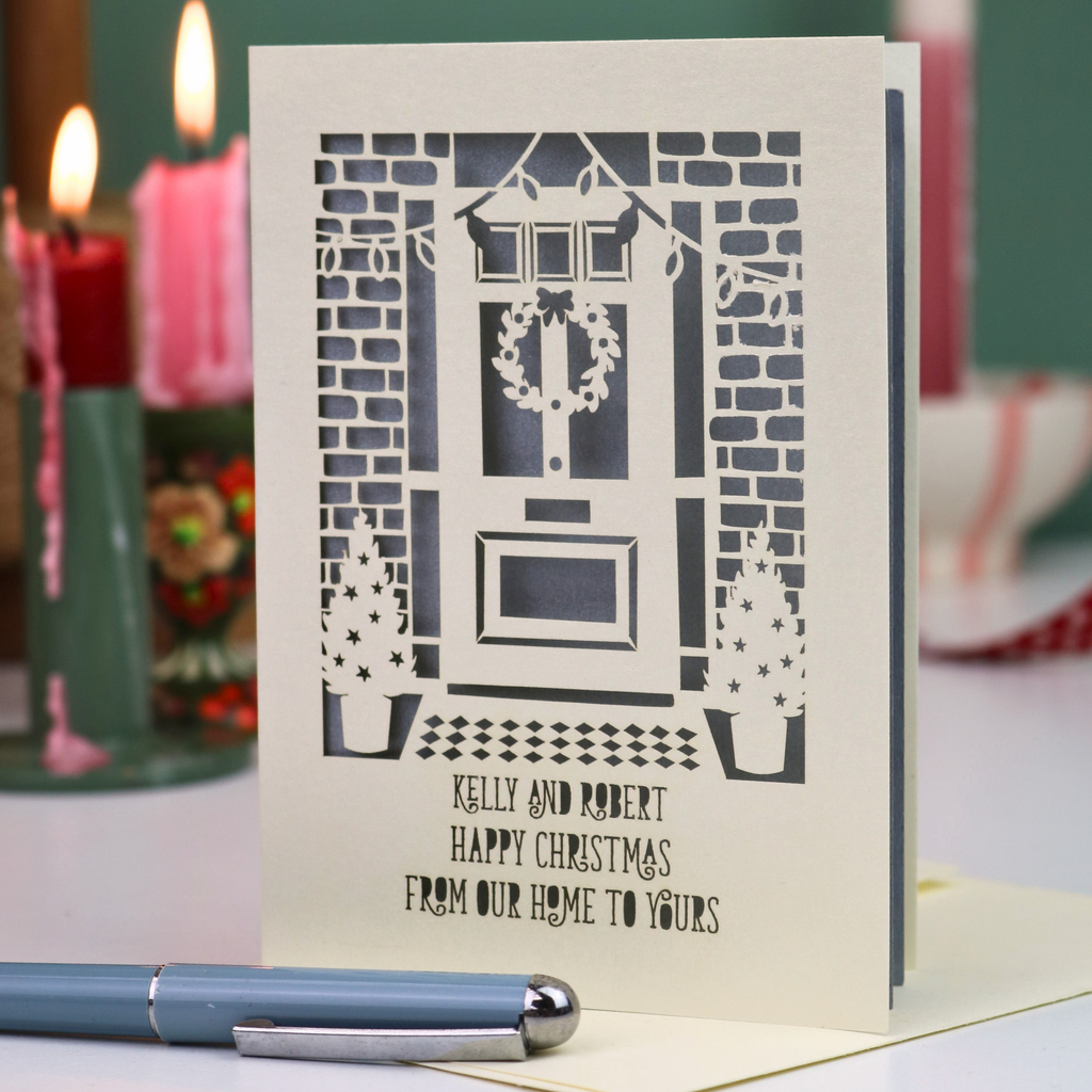 A handmade Christmas greeting card featuring a laser-cut design of a front door with a wreath, brick walls, and star-patterned trees, with the text 'KELLY AND ROBERT HAPPY CHRISTMAS FROM OUR HOME TO YOURS'. The card is displayed on a table with a blue pen in the foreground and blurred red and green candles in the background.