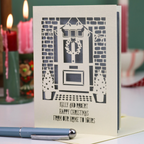 A handmade Christmas greeting card featuring a laser-cut design of a front door with a wreath, brick walls, and star-patterned trees, with the text 'KELLY AND ROBERT HAPPY CHRISTMAS FROM OUR HOME TO YOURS'. The card is displayed on a table with a blue pen in the foreground and blurred red and green candles in the background.