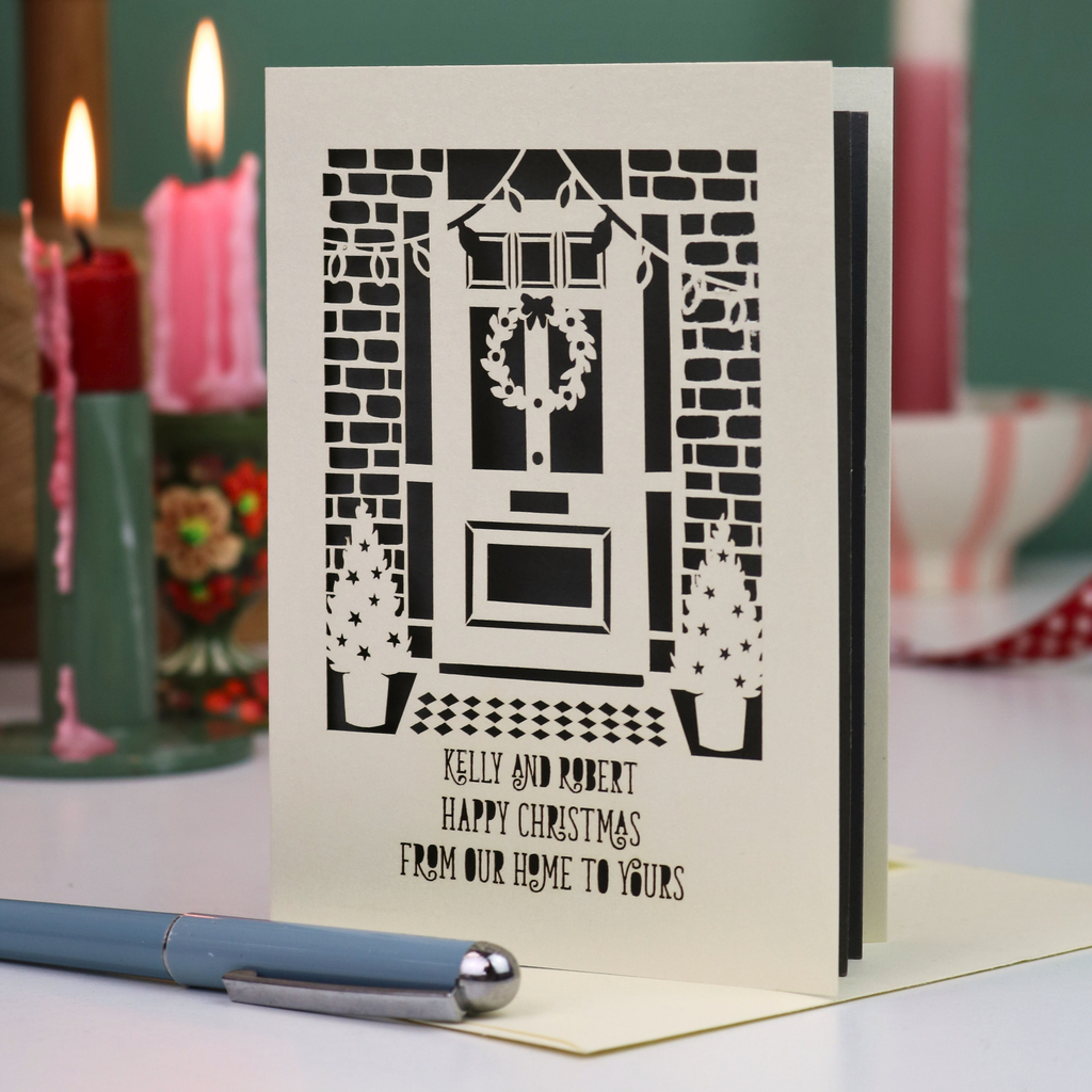 A personalized Christmas greeting card featuring a laser cut illustration of a front door with a wreath, set on a table with a blue pen in the foreground and blurred candles in the background.