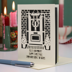 A personalized Christmas greeting card featuring a laser cut illustration of a front door with a wreath, set on a table with a blue pen in the foreground and blurred candles in the background.