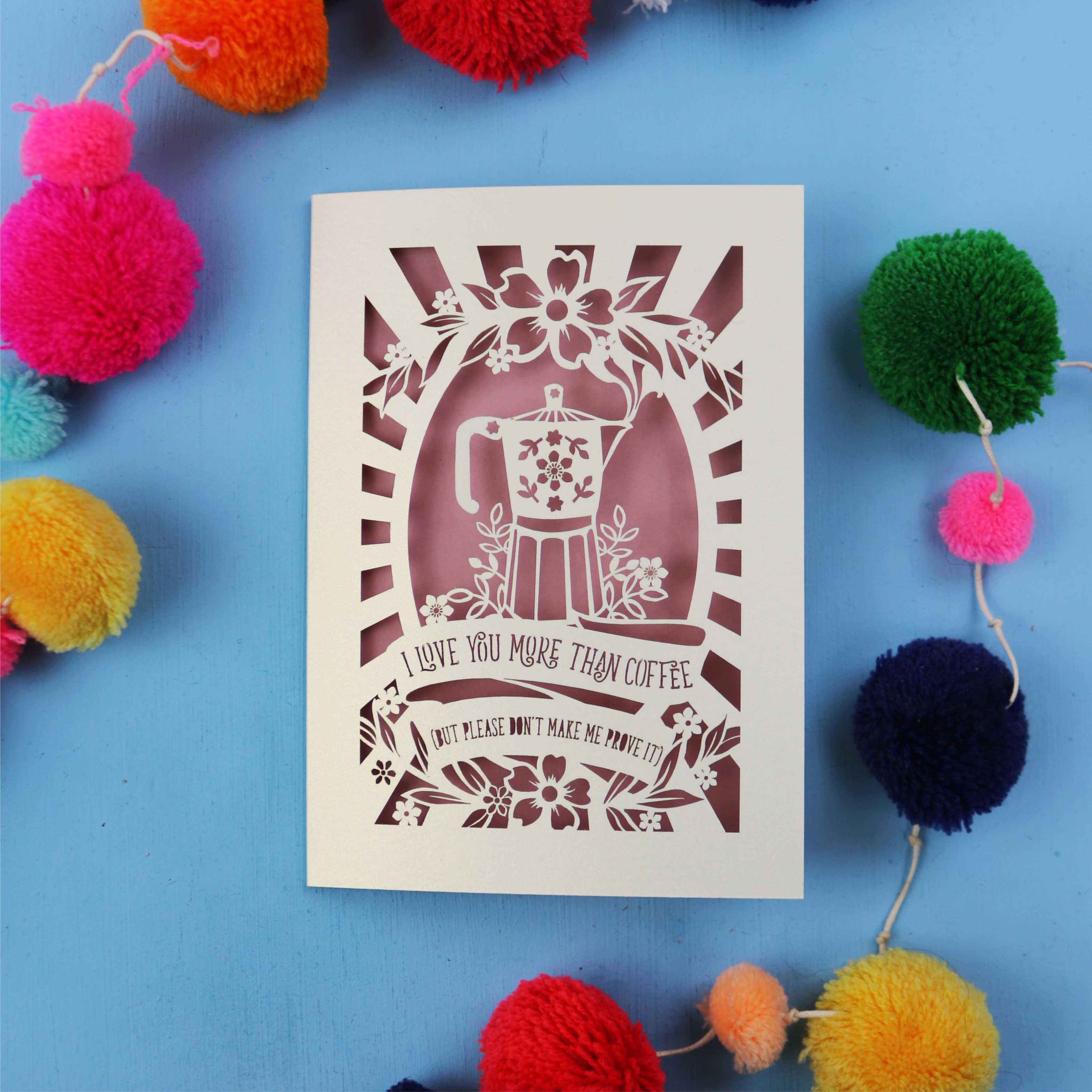 Funny Valentine's card with coffee-themed design and the words "I love you more than coffee, but please don't make me prove it)" on a blue background with colourful pom-poms.