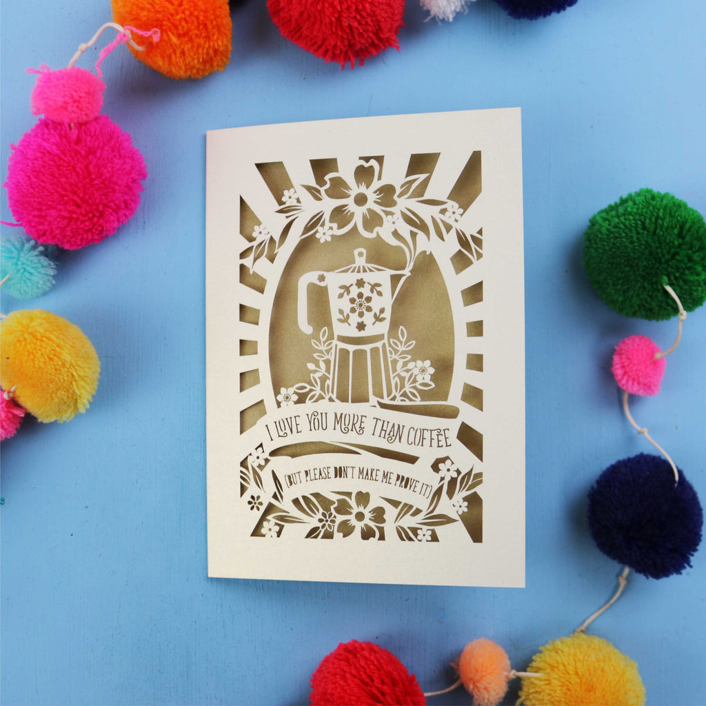 Decorative funny Valentine's card with coffee-themed design and the words "I love you more than coffee, but please don't make me prove it)" on a blue background with colourful pom-poms.