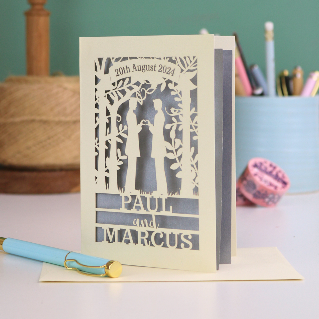 Personalized card with laser-cut design of two men holding hands, on a desk with stationery items.
