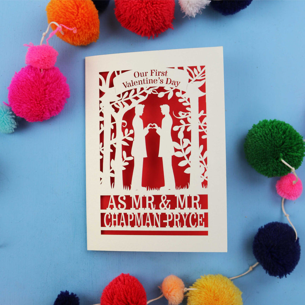 Gay Valentine's Day card with cut-out design of a couple and text, surrounded by colourful pom-poms on a blue background.