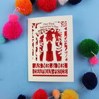 Gay Valentine's Day card with cut-out design of a couple and text, surrounded by colourful pom-poms on a blue background.