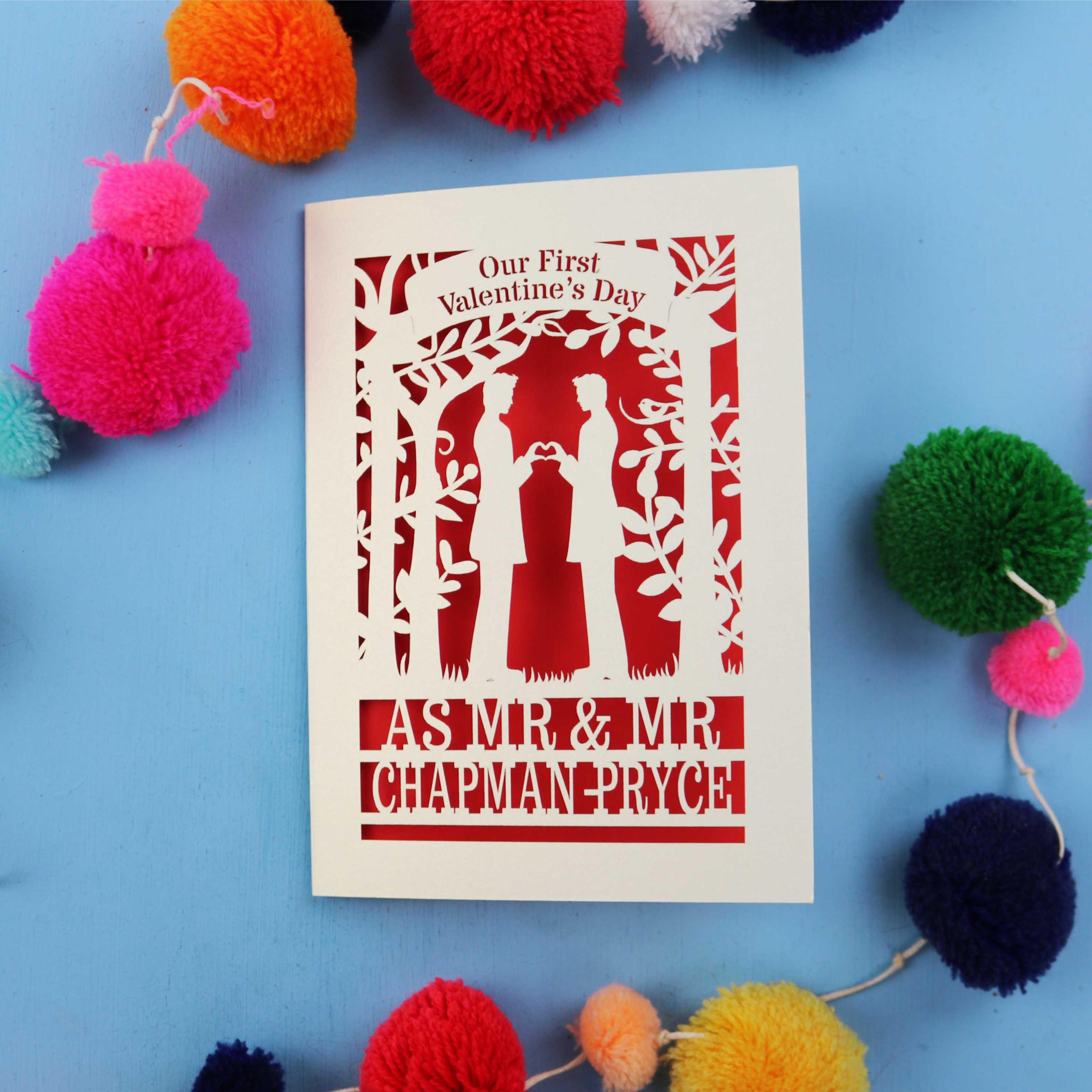 Gay Valentine's Day card with cut-out design of a couple and text, surrounded by colourful pom-poms on a blue background.