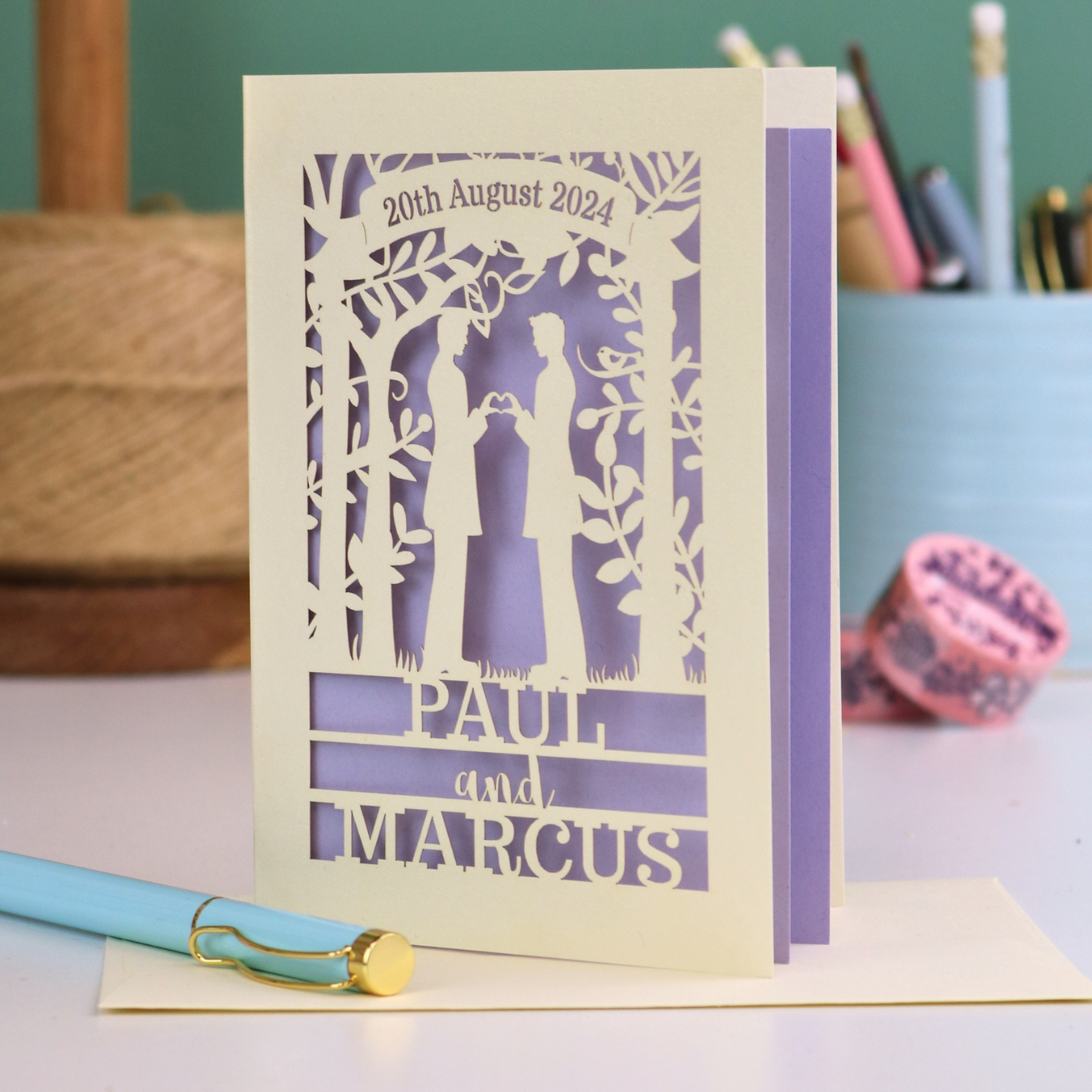 Personalized gey wedding card with laser-cut design and text '20th August 2024, Paul and Marcus' on a desk.