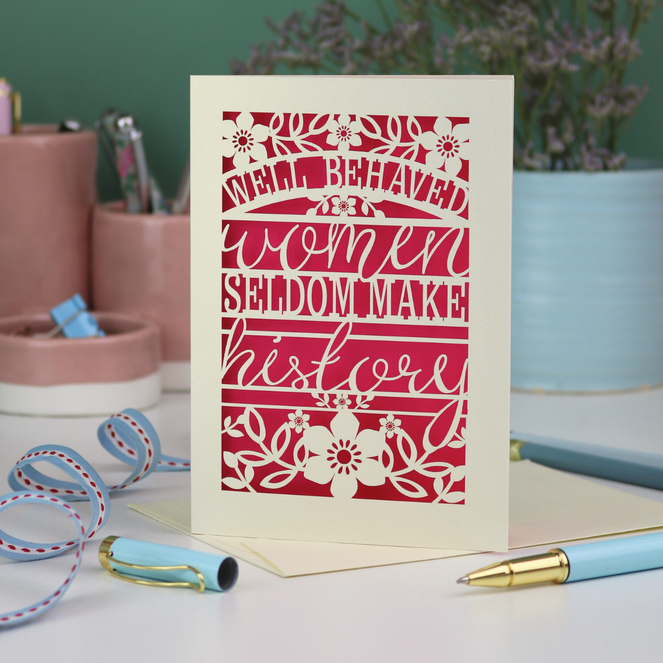 Decorative International Women's Day cut out card with text 'Well Behaved Women Seldom Make History' and floral design on a desk with stationery items.