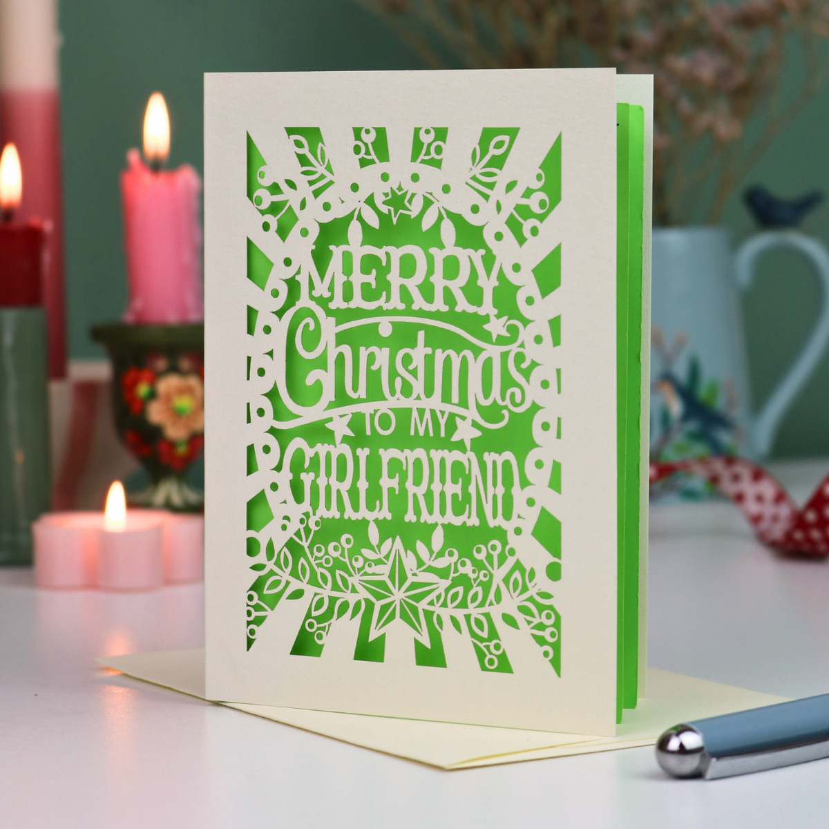 A laser-cut girlfriend Christmas card with the phrase 'Merry Christmas to My Girlfriend' in cut out lettering over a green background, displayed on a table with candles and a pen in a softly lit setting.