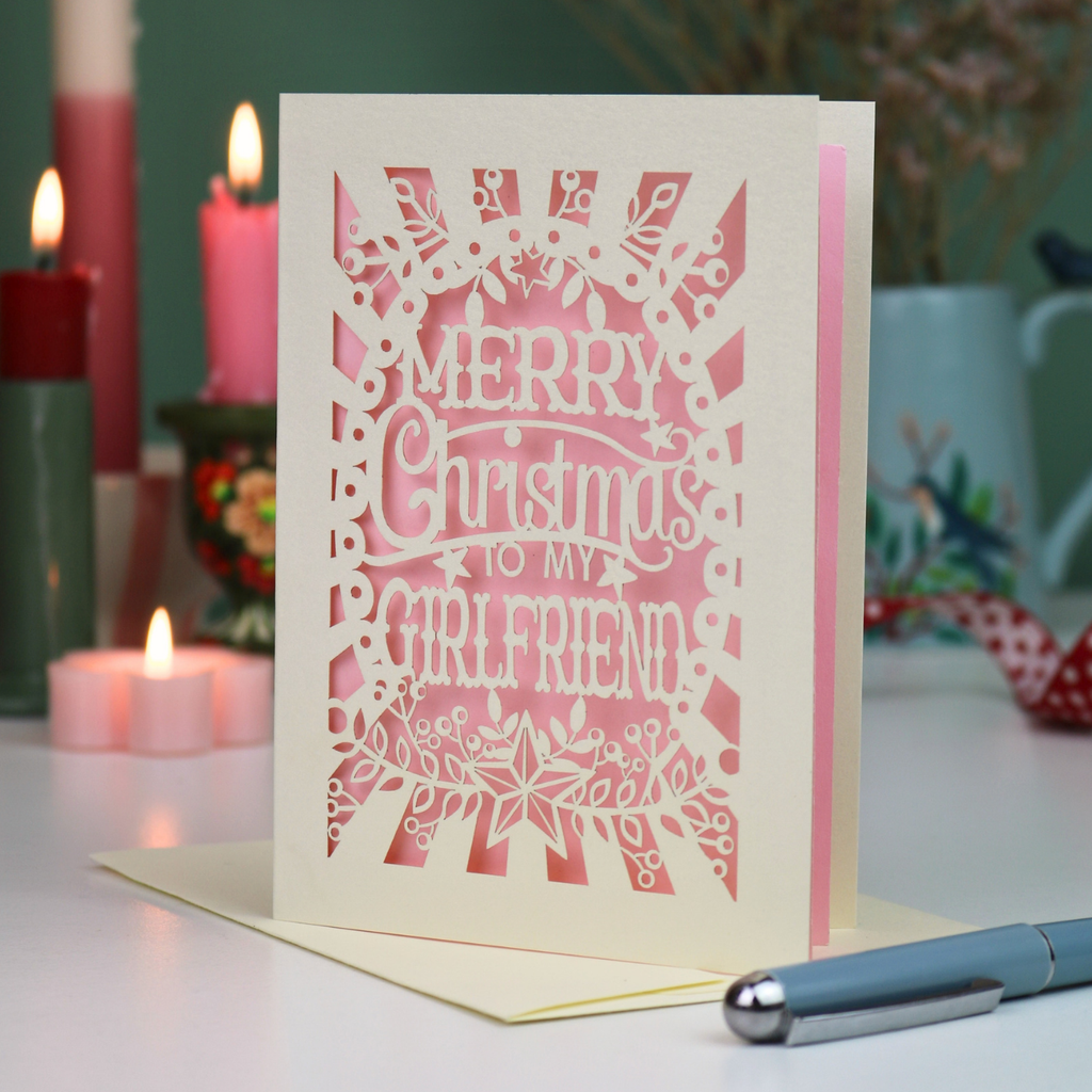 A laser-cut girlfriend Christmas card with the phrase 'Merry Christmas to My Girlfriend' in cut out lettering over a pink background, displayed on a table with candles and a pen in a softly lit setting.