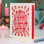 A laser-cut girlfriend Christmas card with the phrase 'Merry Christmas to My Girlfriend' in cut out lettering over a coral background, displayed on a table with candles and a pen in a softly lit setting.