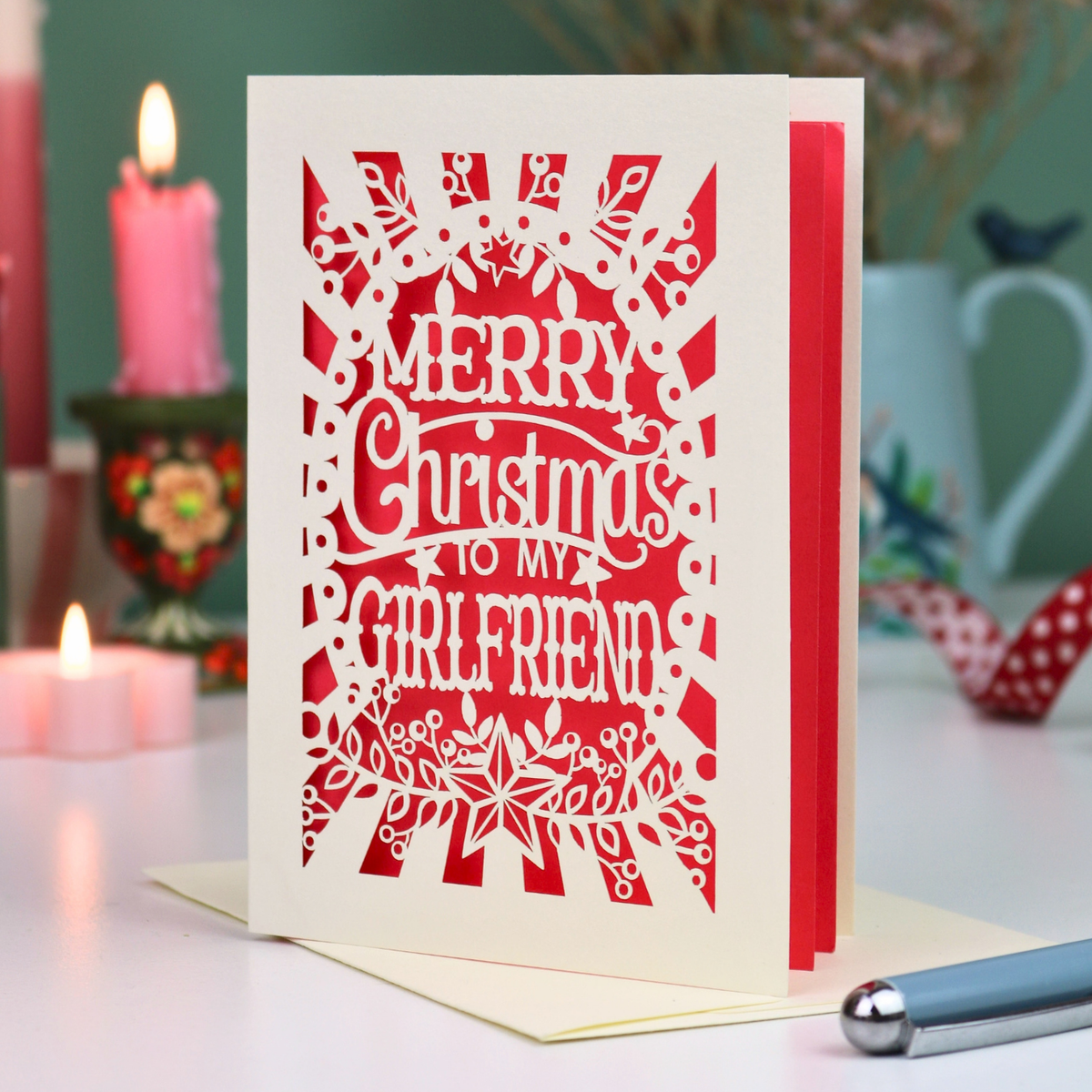 A laser-cut girlfriend Christmas card with the phrase 'Merry Christmas to My Girlfriend' in cut out lettering over a coral background, displayed on a table with candles and a pen in a softly lit setting.