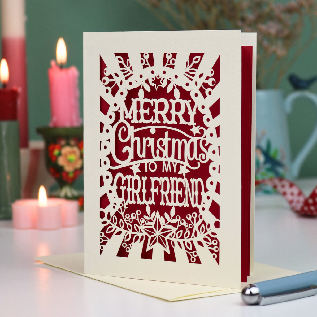 A laser-cut girlfriend Christmas card with the phrase 'Merry Christmas to My Girlfriend' in cut out lettering over a dark red background, displayed on a table with candles and a pen in a softly lit setting.