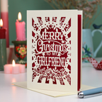 A laser-cut girlfriend Christmas card with the phrase 'Merry Christmas to My Girlfriend' in cut out lettering over a dark red background, displayed on a table with candles and a pen in a softly lit setting.