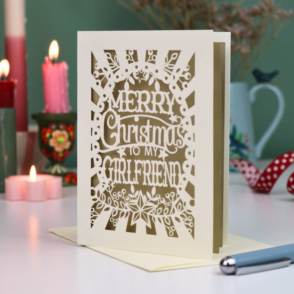 A laser-cut girlfriend Christmas card with the phrase 'Merry Christmas to My Girlfriend' in cut out lettering over a gold leaf background, displayed on a table with candles and a pen in a softly lit setting.