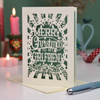 A laser-cut girlfriend Christmas card with the phrase 'Merry Christmas to My Girlfriend' in cut out lettering over a sage background, displayed on a table with candles and a pen in a softly lit setting.