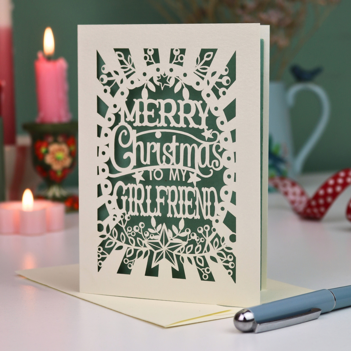 A laser-cut girlfriend Christmas card with the phrase 'Merry Christmas to My Girlfriend' in cut out lettering over a sage background, displayed on a table with candles and a pen in a softly lit setting.