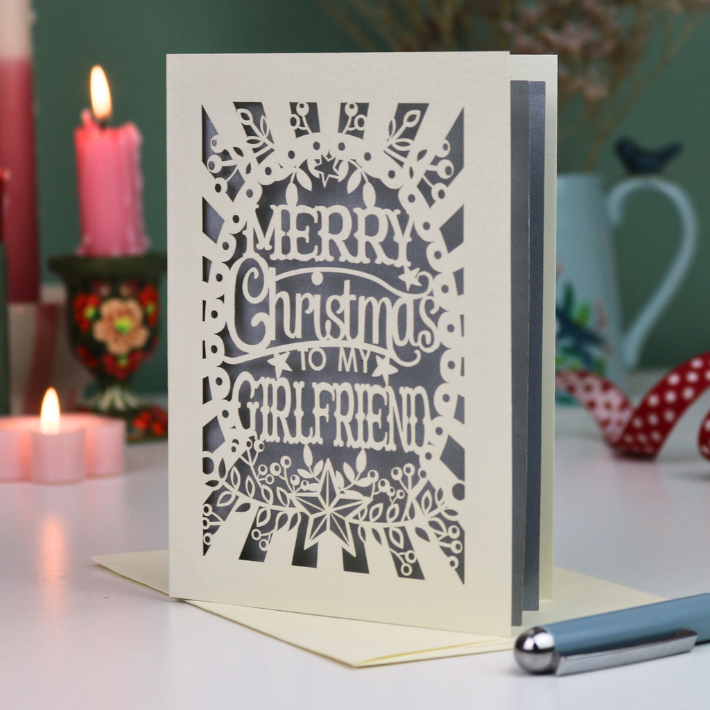 A laser-cut girlfriend Christmas card with the phrase 'Merry Christmas to My Girlfriend' in cut out lettering over a silver background, displayed on a table with candles and a pen in a softly lit setting.