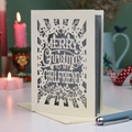 A laser-cut girlfriend Christmas card with the phrase 'Merry Christmas to My Girlfriend' in cut out lettering over a silver background, displayed on a table with candles and a pen in a softly lit setting.