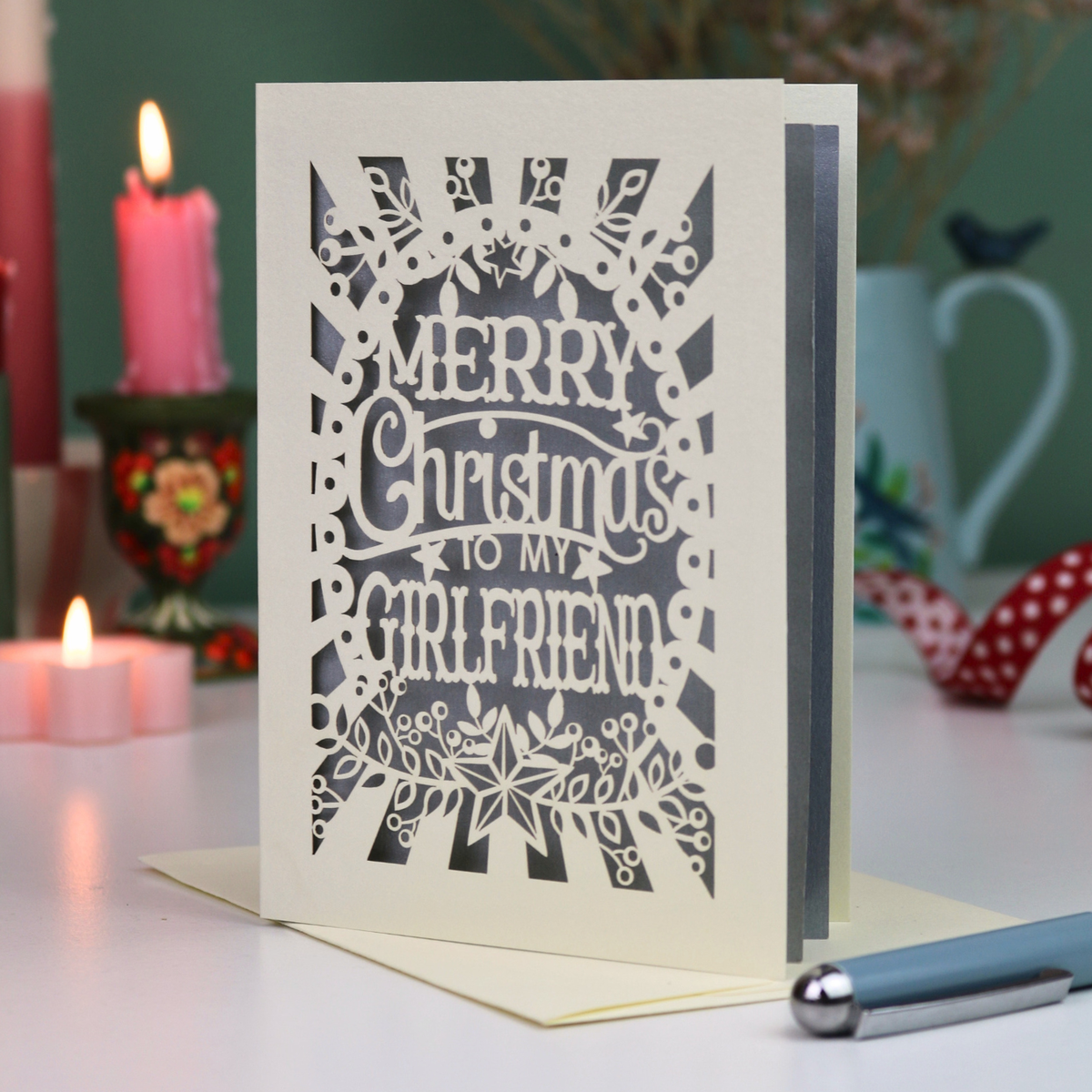 A laser-cut girlfriend Christmas card with the phrase 'Merry Christmas to My Girlfriend' in cut out lettering over a silver background, displayed on a table with candles and a pen in a softly lit setting.