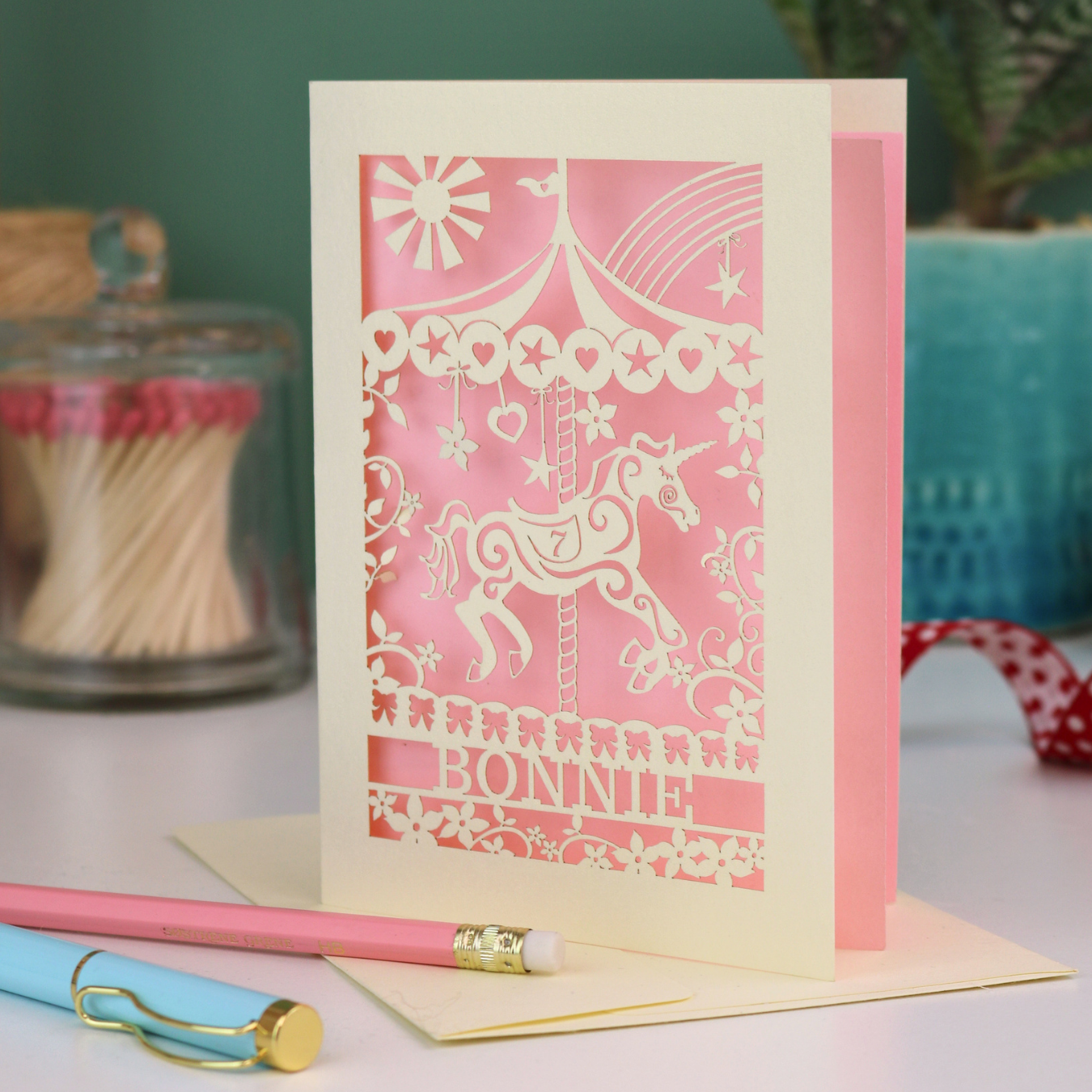Decorative birthday card with cut out unicorn carousel design and 'Bonnie' text on a table with stationery items.