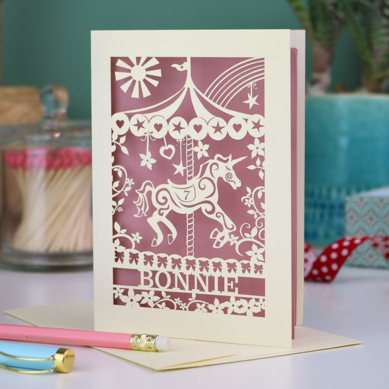 Decorative birthday card with cut out unicorn carousel design and 'Bonnie' text on a table with stationery items.