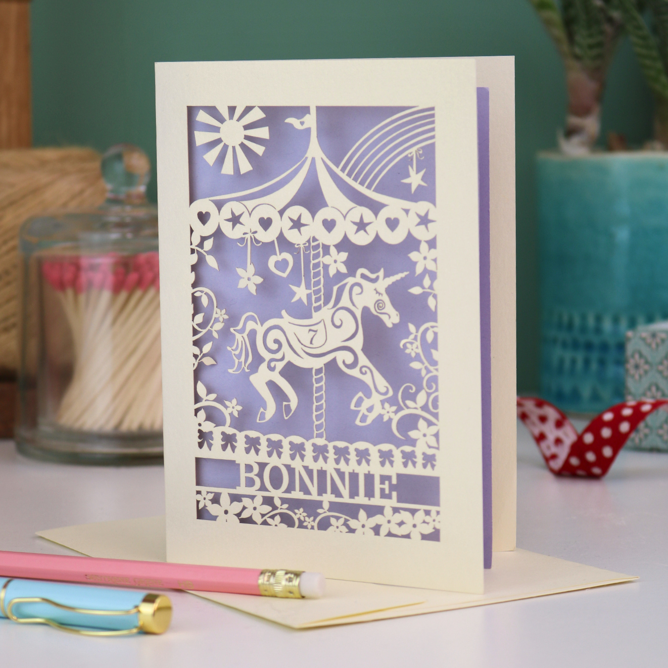 Personalised Laser-cut birthday card with unicorn design and 'Bonnie' text on a table with stationery items.
