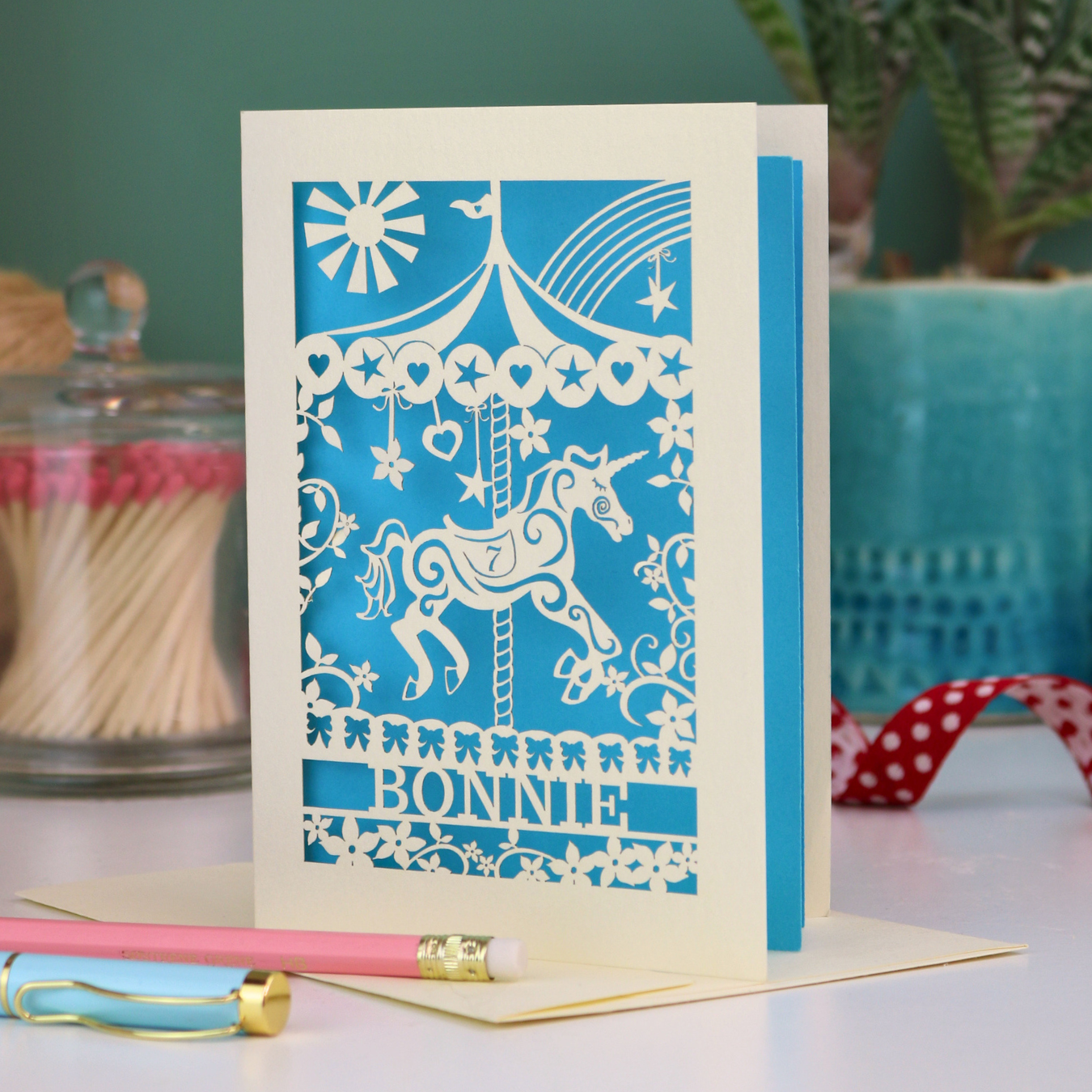 Decorative personalised birthday card with cut out unicorn carousel design and 'Bonnie' text on a table with stationery items.