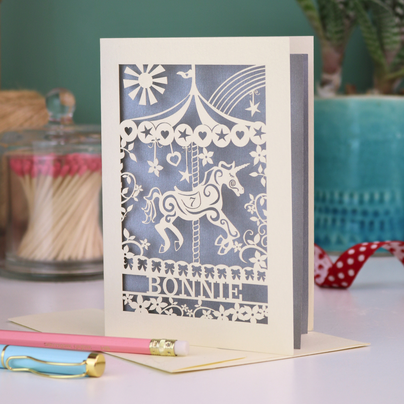Decorative birthday card with cut out unicorn carousel design and 'Bonnie' text on a table with stationery items.