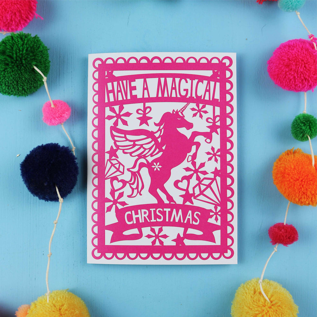 Pink Christmas card with unicorn design on a blue background with colourful pom-poms.