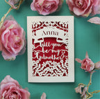 A personalised papercut card, laser cut from cream card and cut away to reveal a pink paper insert. Cut away design shows a banner with recipient's name and text underneath in a script font reads "Will you be my Godmother?"