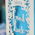 Laser-cut 'Good Luck' card with decorative elements and personalised with the name "Maya" in stencil font on a banner above the text 'Good Luck' in a script font.