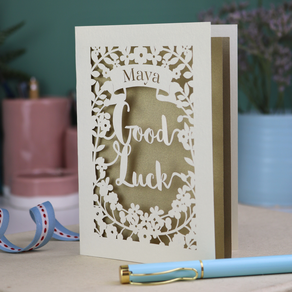 Laser-cut 'Good Luck' card with decorative elements and personalised with the name "Maya" in stencil font on a banner above the text 'Good Luck' in a script font.
