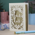 Laser-cut 'Good Luck' card with decorative elements and personalised with the name "Maya" in stencil font on a banner above the text 'Good Luck' in a script font.