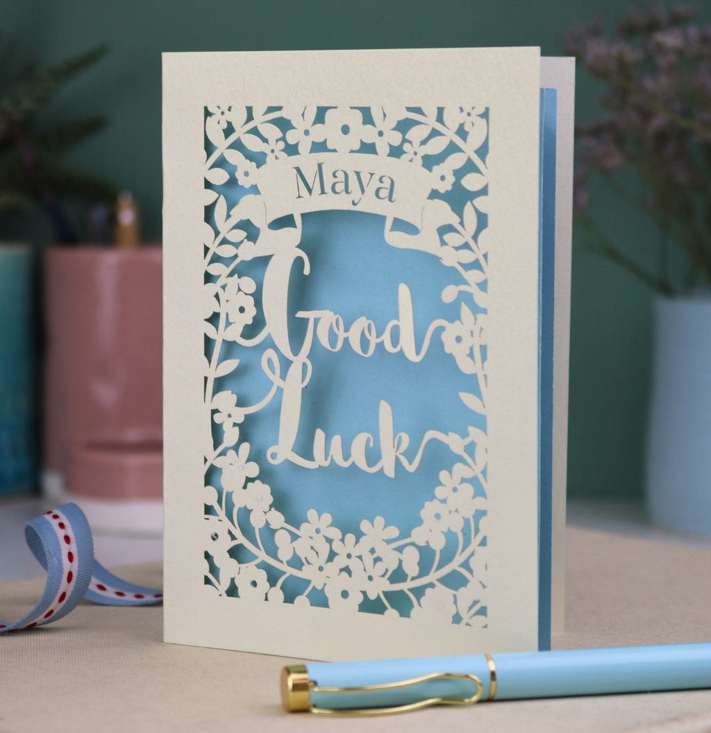 Laser-cut 'Good Luck' card with decorative elements and personalised with the name "Maya" in stencil font on a banner above the text 'Good Luck' in a script font.