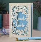 Laser-cut 'Good Luck' card with decorative elements and personalised with the name "Maya" in stencil font on a banner above the text 'Good Luck' in a script font.