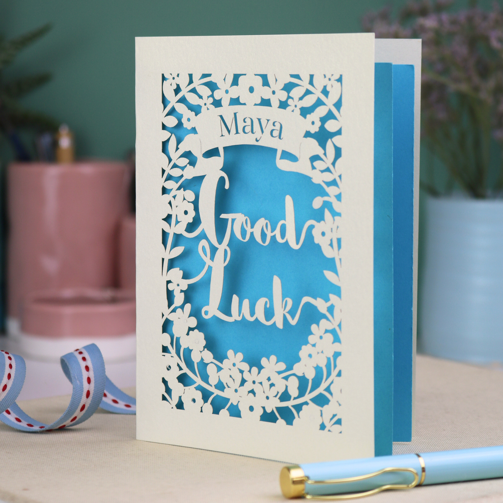 Laser-cut 'Good Luck' card with decorative elements and personalised with the name "Maya" in stencil font on a banner above the text 'Good Luck' in a script font.