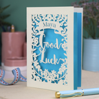 Laser-cut 'Good Luck' card with decorative elements and personalised with the name "Maya" in stencil font on a banner above the text 'Good Luck' in a script font.