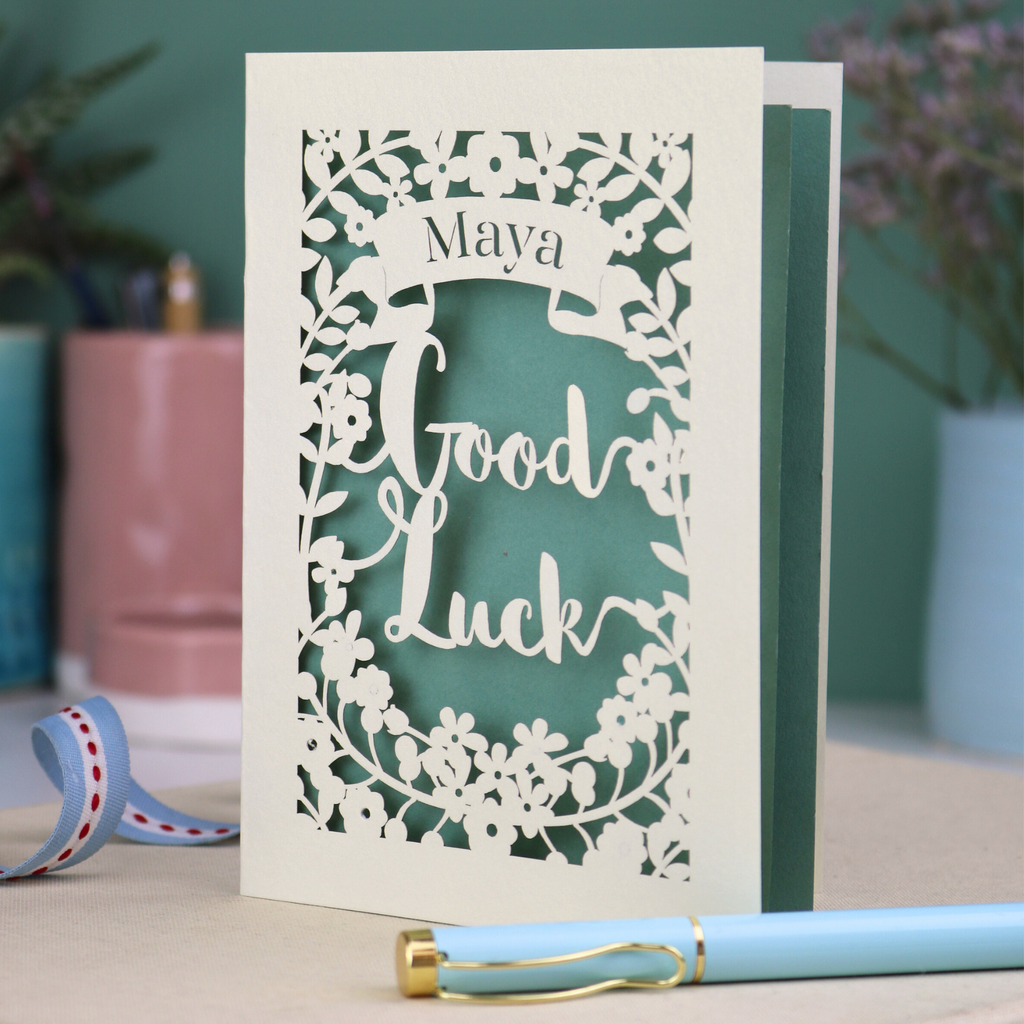 Laser-cut 'Good Luck' card with decorative elements and personalised with the name "Maya" in stencil font on a banner above the text 'Good Luck' in a script font.