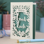 Laser-cut 'Good Luck' card with decorative elements and personalised with the name "Maya" in stencil font on a banner above the text 'Good Luck' in a script font.