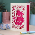 Laser-cut 'Good Luck' card with decorative elements and personalised with the name "Maya" in stencil font on a banner above the text 'Good Luck' in a script font.