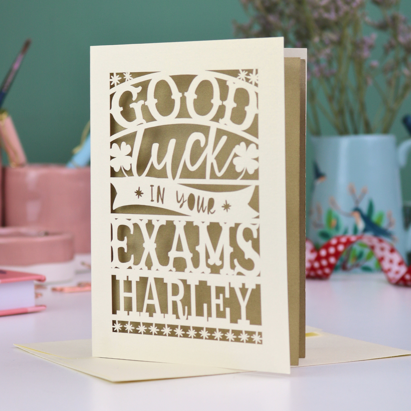 Personalised Laser-cut 'Good luck in your exams' card on a desk with stationery items.