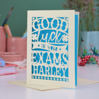 Personalized good luck card for exams with blue text on a light background, placed on a desk with stationery items.