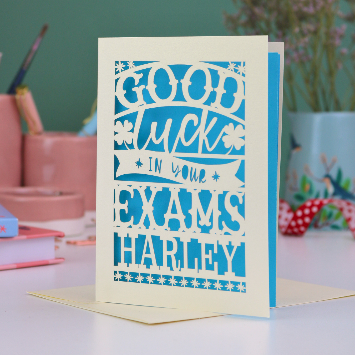 Personalized good luck card for exams with blue text on a light background, placed on a desk with stationery items.