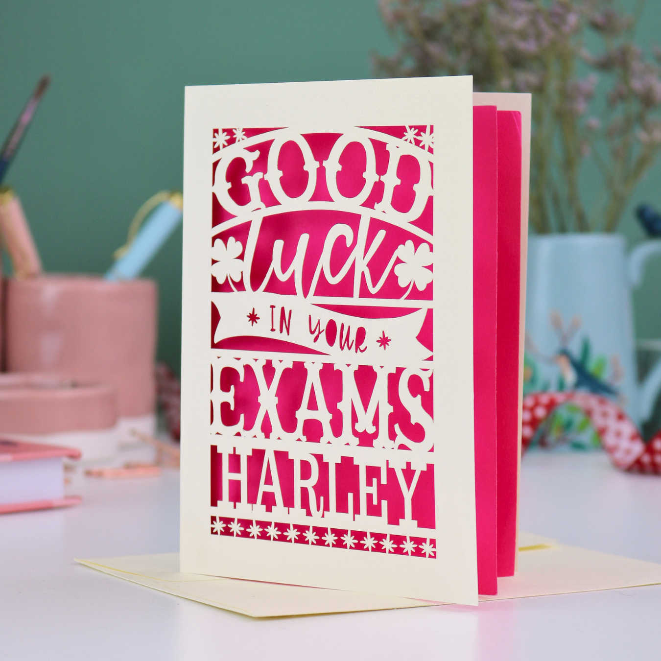 Greeting card with 'Good luck in your exams Harley' text on a desk with stationery items.
