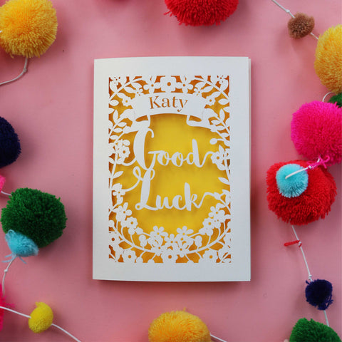 Laser cut personalised Good Luck card. Cut from cream card with a bright yellow background. Shows the words good luck surrounded with flowers and a banner for the name.