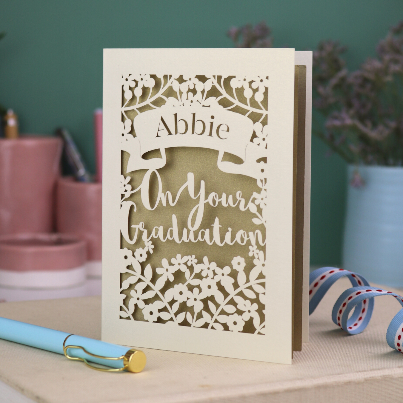 Laser cut Graduation card with personalized name 'Abbie' and floral cut out design, on a table with decorative elements.