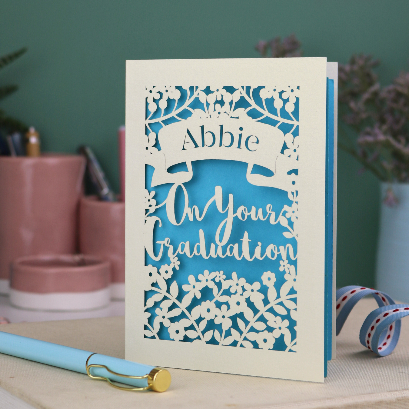 Laser cut Graduation card with personalized name 'Abbie' and floral cut out design, on a table with decorative elements.